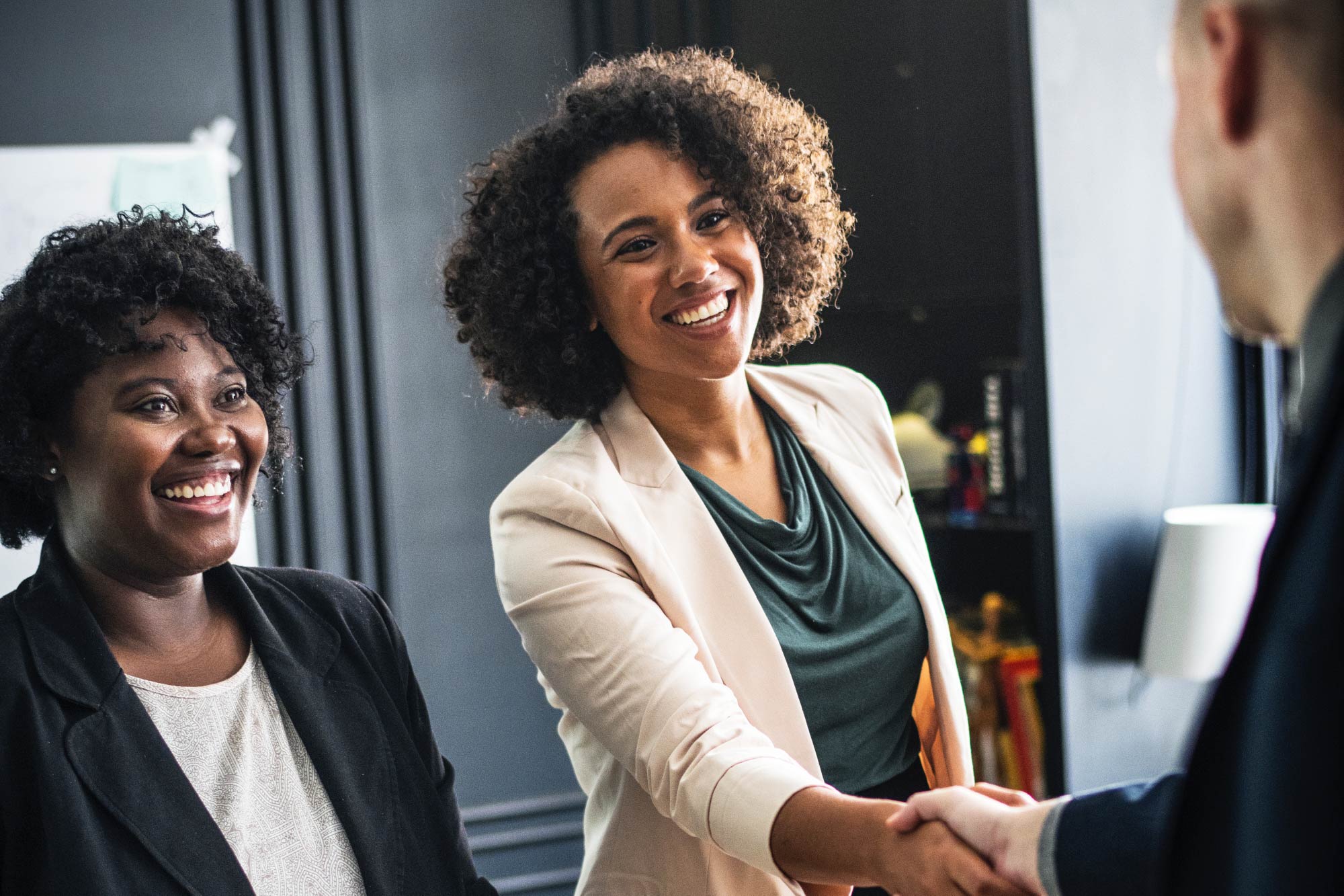 Two black women smiling and greeting someone in an office. One woman is shaking hands with a white man.
